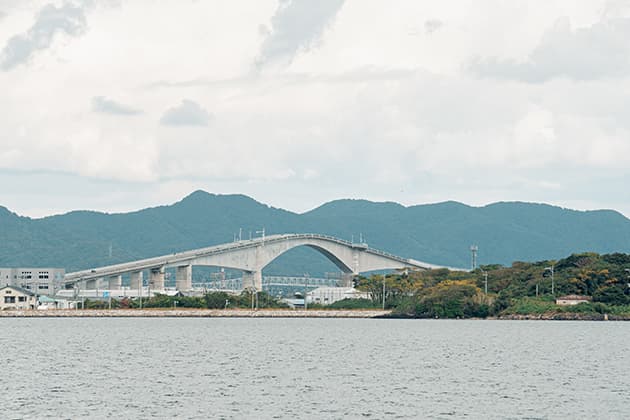江島大橋 風景 えしまおおはし テレビCMで一躍有名になり「ベタ踏み坂」と名付けられた坂。正式名は「江島大橋(えしまおおはし)」。島根県松江市八束町から鳥取県境港市渡町へ中海(なかうみ)をまたいで結ぶ日本一のPCラーメン構造の橋。豪華クルーズ船並の船が潜れるよう最上部は高さ約45mにもなります。松江市側の勾配が高く、橋の横から見るとその形状がよくわかります。鳥取県境港市