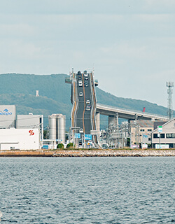 江島大橋 正面 えしまおおはし テレビCMで一躍有名になり「ベタ踏み坂」と名付けられた坂。正式名は「江島大橋(えしまおおはし)」。島根県松江市八束町から鳥取県境港市渡町へ中海(なかうみ)をまたいで結ぶ日本一のPCラーメン構造の橋。豪華クルーズ船並の船が潜れるよう最上部は高さ約45mにもなります。松江市側の勾配が高く、橋の横から見るとその形状がよくわかります。鳥取県境港市