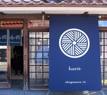 赤瓦五号館 のれん店舗外観　久楽アカガワラゴゴウカン クラ白壁⼟蔵群にある、築100年の古民家をDIYでリノベーションした古民家カフェ。店内からは伝統的な建造群の趣ある風景が楽しめます。石臼で挽いたコーヒーに、砂糖の代わりに小豆を入れて味わう伝統の「石臼珈琲」をはじめ、まちを散策しながら飲み歩きができる「クラボトル」や四季折々の「くらだんご」がおすすめ⿃取県倉吉市新町